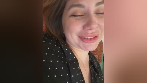 Video of a young woman with fair skin, brown hair, and a nose ring, wearing a black polka-dotted blouse, smiling with closed eyes, against a blurred background.