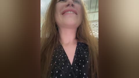 A close-up video of a smiling, fair-skinned woman with long, straight, light brown hair, wearing a black dress with white polka dots, taken from a low angle.