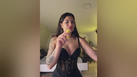 Video of a Latina woman with long dark hair, wearing a black lace lingerie top, holding a yellow flower near her lips in a dimly lit bedroom.