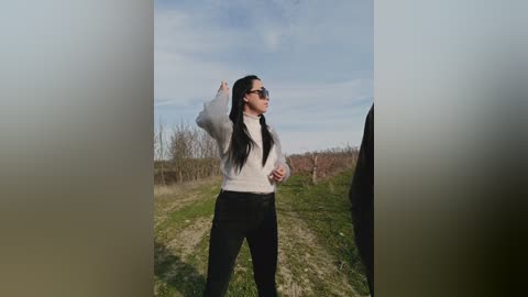 Video of an Asian woman with long black hair, wearing sunglasses and a white sweater, gesturing with her right hand while standing on a dirt path in a grassy field under a partly cloudy sky.