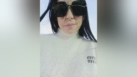 Video of a woman with fair skin, long black hair, and black sunglasses, standing with her head slightly tilted. She wears a white, high-neck turtleneck with black text on the right sleeve. Background is blurred.