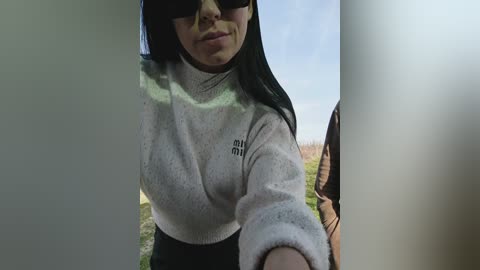 Video of a woman with long black hair, wearing sunglasses and a white turtleneck sweater, standing in front of a blurred outdoor landscape.