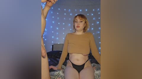 Video of a young woman with light skin, short blonde hair, wearing a cropped brown sweater and black thong, sitting on a bed with a floral-patterned blanket. A man's hand is visible reaching into the frame from the left.