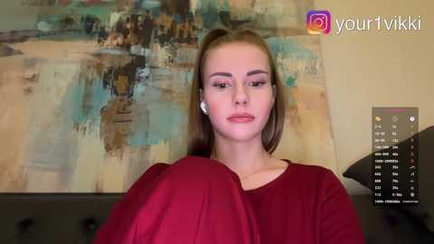 Video of a young Caucasian woman with fair skin, light brown hair, and a slim physique, wearing a red blouse, standing indoors with abstract art in the background and a smartphone displaying a virtual makeup app.