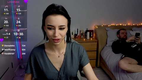Video of a woman with fair skin and dark hair, wearing a grey ribbed top, sitting on a bed in a dimly lit bedroom. Background shows a man sleeping.