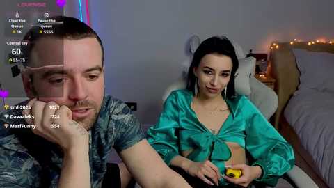 Video of a man with a beard in a camo shirt, and a woman in a green satin crop top, sitting on a bed in a dimly lit room.