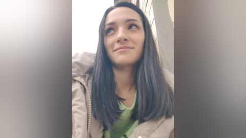 Video of a young woman with long, straight, dark hair, wearing a beige jacket over a green sweater, looking upward thoughtfully. Background is blurred.