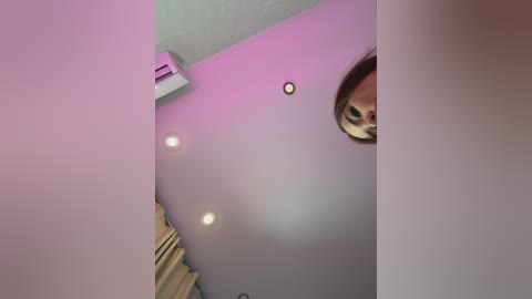 Video of a ceiling with three recessed lights, a white air conditioner, and a small, blurred person peeking around a corner, partially visible. The ceiling is painted light gray, and the room has beige curtains.