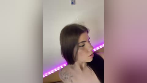 Video of a young woman with long dark hair, light skin, wearing a white top, standing against a plain white wall with purple LED lights.