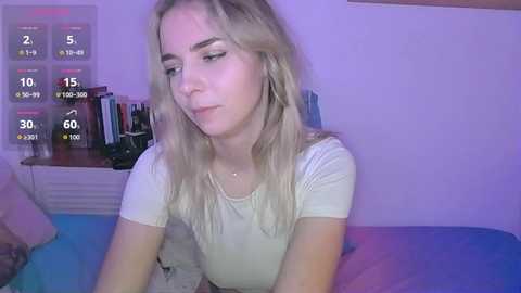 Video of a young, fair-skinned woman with blonde hair in a casual beige top, sitting on a bed with blue and purple bedding, in a softly lit room with a digital clock showing 2:59 AM.