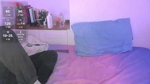 Video of a bedroom with a light blue pillow and a dark green blanket on a bed, beside a white dresser with books and a lamp.