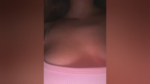 Video of a woman's bare shoulder and chest, wearing a light pink, ribbed tank top, with a blurred background of reddish-brown hues.