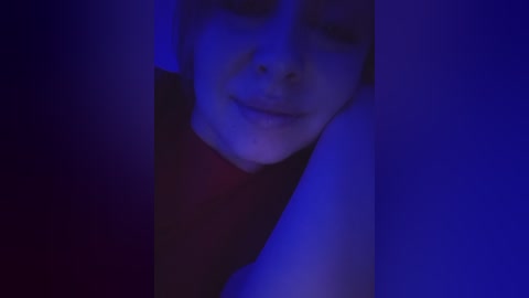 A dimly lit video featuring a young person with fair skin and short brown hair, lying on their side with a gentle smile, under blue lighting.