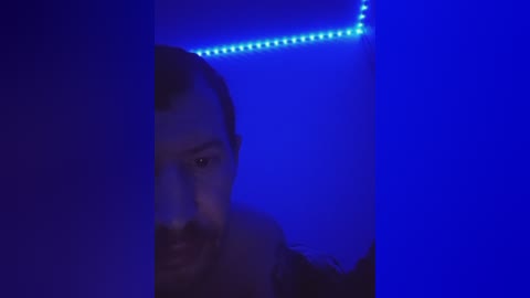 Video of a man with short dark hair, beard, and glasses, partially obscured by a blue light, looking intense, in a dimly lit room.