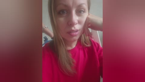 Video of a Caucasian woman with long, straight, light brown hair, wearing a bright red shirt, with blurred, red-tinted vertical edges, and a blurred background.