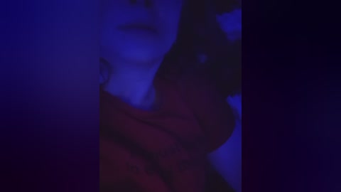 A dimly-lit video features a close-up of a woman's face, partially obscured by shadows. She has a fair complexion, with a hint of red lipstick and a subtle smile. The background is a deep blue, creating a moody, intimate atmosphere.