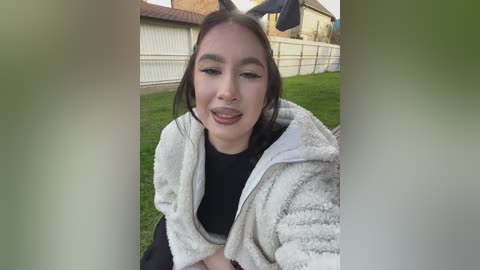 Video of a young woman with light brown skin, wearing a fluffy white jacket, black top, and bunny ears, sitting on a green lawn with a white fence and house background.
