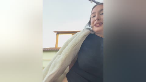 Video of a smiling Asian woman with dark hair, wearing a black top, standing in a bright room with a beige towel draped over her shoulder.