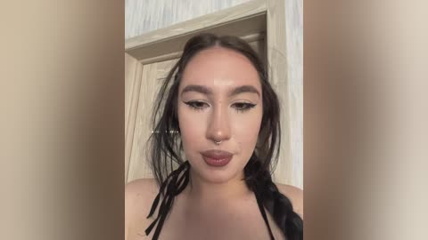 Video of a young woman with light skin, dark braided hair, and heavy makeup, including dark eyeliner and nude lipstick, standing in front of a beige door.