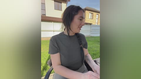 A video of a young woman with long, dark hair in a braid, wearing a grey t-shirt and sitting on a wheelchair in a suburban backyard.