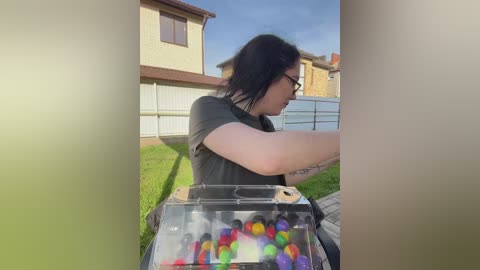 Video of a woman with glasses, black hair, and tattoos on her arm, wearing a dark shirt, playing with colorful beads on a small table outdoors.