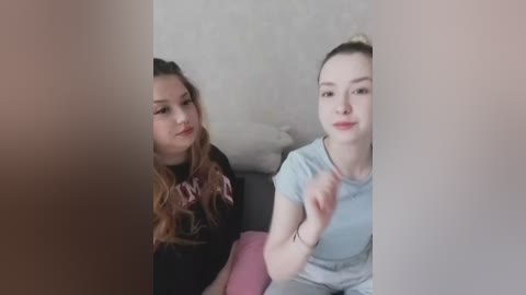 Video of two young women seated side by side, one with long wavy brown hair, wearing a black sweater, and the other with straight brown hair, dressed in a light gray t-shirt. They are indoors with a beige wallpaper background.
