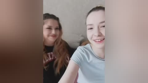 A video of two young girls with light skin, smiling. The girl in the foreground wears a light blue shirt, while the girl behind has long brown hair, wearing a black shirt with a pattern.