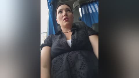 Video of a woman with fair skin and dark hair, wearing a black lace dress, seated in a car with blue curtains.