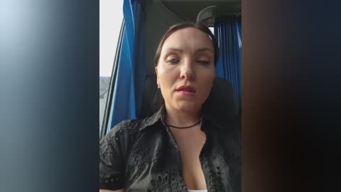 Video of a middle-aged woman with light skin and dark hair, wearing a black lace blouse and black choker necklace, sitting in a bus with blue curtains.