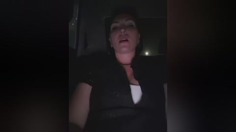 A dimly lit video shows a woman with short dark hair, wearing a black top and white shirt, looking startled and slightly out of focus, possibly inside a car at night.
