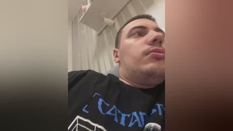 Video of a young man with short dark hair and light skin, wearing a black t-shirt with \"KAPATID\" in blue text. He's making a pouty face and leaning against a beige wall with a ceiling fan above.