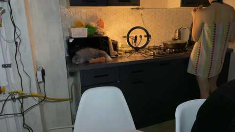 Video of a cozy kitchen with a black countertop, a grey cat sleeping on a mat, a ring light, a striped apron, and white chairs.