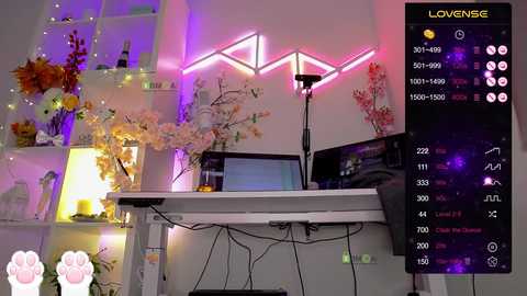 Video of a cozy, modern home office with white walls, pink neon lights, and a white desk. A large monitor displays a camera feed, a lamp, and a vase with flowers. A \"LOVENSE\" screen displays stats.