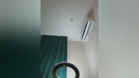 Video of a minimalist, modern room with a white ceiling, light beige walls, and a teal curtain on the left. An air conditioner is mounted on the wall, and a circular, black-framed mirror hangs below it.