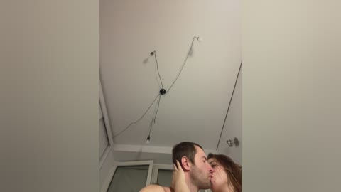 Video of a man and woman kissing in a corner of a room with white walls, a ceiling fan, and a window with blinds partially open.