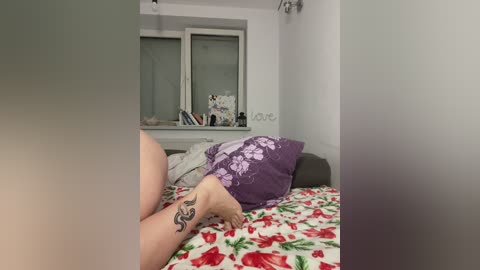 Video of a person's bare legs with a tattoo on the left ankle, lying on a bed with a red and green floral duvet, in a modestly decorated room with white walls and a window.