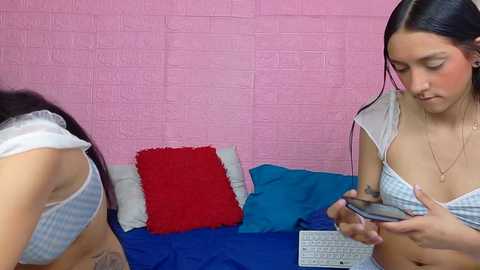 Video of two young women with medium skin tones, one holding a camera, the other in a blue and white checkered bra, lying on a bed with red and blue pillows against a pink brick wall.