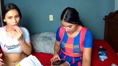 Video of two young women, one with a football-themed crop top and the other in a Barcelona jersey, eating chips on a bed with red sheets in a casual indoor setting.