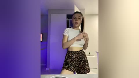 Video of a young woman with fair skin, dark hair, wearing a white crop top and leopard-print shorts, posing in a dimly lit room with a wall-mounted phone.