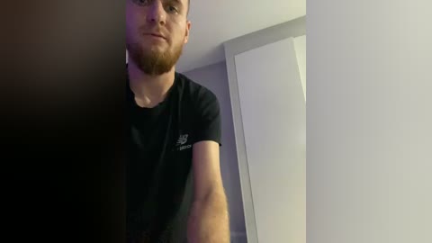 A video of a young, fair-skinned man with a short beard, wearing a black t-shirt, standing indoors with a gray wall and door in the background.