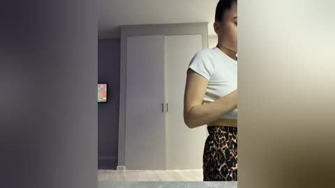 Video of a woman with short black hair, wearing a white crop top and leopard-print skirt, standing in a modern, dimly lit room with white walls and a glass door.