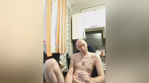 Video of a bald, fair-skinned, shirtless man with a thin physique sitting on a kitchen counter in a small, cluttered kitchen with white cabinets, orange curtains, and a dark countertop.