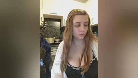 A candid video of a young woman with long, straight auburn hair, wearing a low-cut black dress with lace trim, standing in a modern kitchen with white cabinets and a dark backsplash.