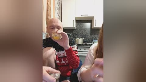 Video of a bald, middle-aged man in a red sweater with Nordic patterns, drinking orange juice in a modern kitchen.
