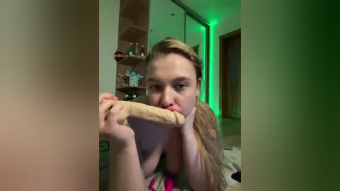 A video of a young woman with light skin, blonde hair, and average-sized breasts, licking a wooden dildo while lying on a bed in a dimly lit, green-lit room.
