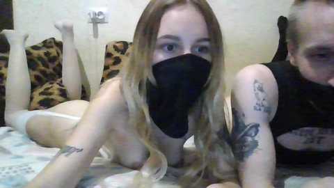 Video of a young, fair-skinned woman with long blonde hair and a black face mask, lying on a bed with leopard-print pillows. She is topless, revealing a tattoo on her arm.
