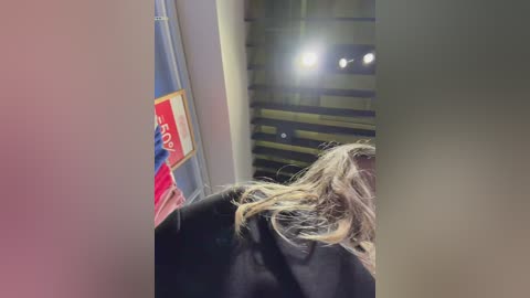 Video of a woman with shoulder-length blonde hair, wearing a black jacket, standing indoors. Background features a red poster and a bright light source creating a dramatic contrast.