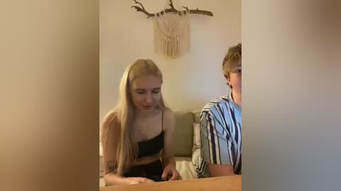 Video of a young blonde woman with long hair in a black top, leaning forward, and a young boy with short hair in a striped shirt, sitting on a beige couch in a cozy room with a macram\u00e9 wall hanging.