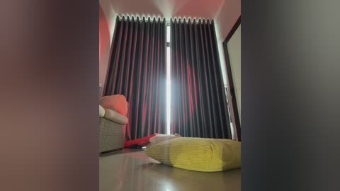 A video of a modern living room with a large window adorned with dark, floor-length curtains. A wicker armchair and a yellow throw pillow are situated in the foreground, casting soft shadows. The room features neutral tones and a minimalist design.