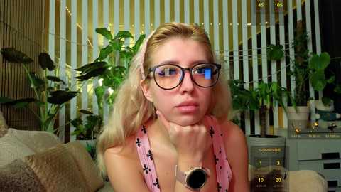 Video of a blonde woman with glasses and a pink dress, surrounded by green plants in a cozy, dimly lit room.